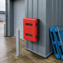 Red double fire extinguisher cabinet wall mounted on grey profiled steel cladding outside an industrial unit, beside a roller shutter door, with a galvanised bollard on damp concrete and blue plastic pallets stacked to the right