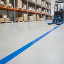 Blue PVC floor marking tape line applied on a warehouse floor marking an equipment zone, with racking and a pallet truck in the background