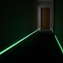Photoluminescent egress marking tape glowing along a dark corridor skirting board