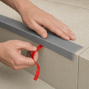 Hand peeling the red adhesive liner and pressing a grey PVC stair nosing onto a stone step edge