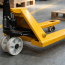 Close-up of the hand pallet truck running gear on a warehouse floor showing the white nylon steering wheels, hydraulic pump with coil spring and chain linkage and the nylon tandem load roller under the yellow fork