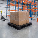Black recycled plastic pallet loaded with a shrink-wrapped stack of cardboard cartons on a warehouse floor, with blue and orange pallet racking and a yellow hand pallet truck behind