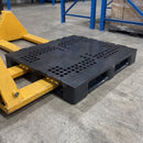 Close-up of the black plastic pallet's perforated deck grid and runner base with the fork of a yellow hand pallet truck alongside on a concrete warehouse floor