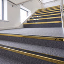 Heavy duty rake back aluminium stair nosing with black insert in mirrorflect gold finish fitted on a stair with grey contract carpet