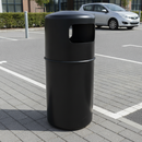 Black 90 litre hooded top litter bin on block paving at the edge of a retail park car park, next to white bay markings, a silver parked car and a steel bollard with a clipped hedge behind