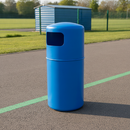 Blue 90 litre hooded top litter bin on tarmac at the edge of a school playground, with a green painted line marking, mesh railing fence, blue storage shed and grass playing field in the background