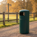 Dark green 90 litre hooded top litter bin on a gravel footpath in a country park, beside a timber post-and-rail fence and a wooden signpost, with autumn trees behind in morning light
