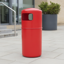 Red 90 litre hooded top litter bin on light grey paving at a leisure centre entrance, next to a stainless steel handrail, glazed doors and a dark grey planter with shrubs