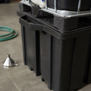 Close-up of the black IBC spill pallet's moulded rim, integral drip tray and perforated grid deck with the base of an IBC cage resting on it, a steel funnel and green transfer hose on the concrete floor alongside