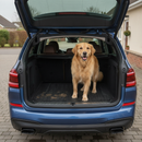 Golden retriever dog standing on a black non-slip rubber car boot liner mat in the open boot of an estate car, with muddy paw prints visible on the mat