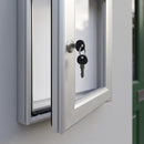 Close-up of the noticeboard door held ajar, showing the satin silver key barrel lock with two black keys, the aluminium extrusion edge, the black rubber weather seal and the white magnetic back panel