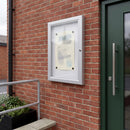 Lockable outdoor noticeboard wall mounted in portrait on a red brick wall beside a dark green entrance door, holding a pale notice behind clear acrylic glazing, with a galvanised handrail and a black planter below