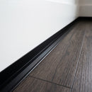 Black anodised aluminium internal corner trim fitted where a dark oak-effect LVT floor meets a white wall in an apartment hallway