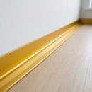 Gold anodised aluminium internal corner trim fitted where a pale oak-effect LVT floor meets a white wall in a hotel corridor