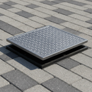 Galvanised steel manhole cover and frame installed in a block-paved driveway, cover lifted slightly to reveal the black polypropylene frame beneath