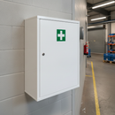 White metal first aid wall cabinet mounted at chest height on a grey block wall in a factory corridor, with a red fire extinguisher, blue stillages and a yellow walkway line beyond