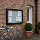 Landscape black weatherproof notice board wall mounted on the red brick wall of a community centre beside a bicycle stand and terracotta planters, blank notices pinned to the grey felt panel