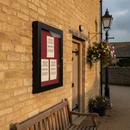 Black outdoor poster case notice board mounted on the Cotswold stone wall of a village hall above a wooden bench, showing blank notices pinned to the red felt backboard