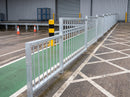 Run of galvanised pedestrian guardrail panels installed to separate a green-painted pedestrian walkway from the vehicle lane in a warehouse service yard, with a yellow and black bollard, orange traffic cones and a loading dock roller shutter behind