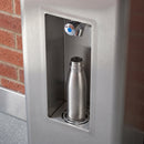 Close-up of the recessed bottle filling alcove on the stainless steel drinking fountain, showing the chromed push-button filler tap with blue index button, the round drain grate and the brushed satin steel grain, with a stainless bottle in place