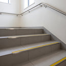 Retro-fit stainless steel stair nosing with CG Yellow RAL 1021 R11 grit tread on a grey tiled public building stairwell