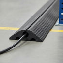 Close-up of the cut end of the rubber cable ramp on a workshop floor, showing the 20mm cable bore, the top entry slot and the tapered ribbed edges with a power cable fitted inside