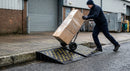Pair of black and yellow rubber kerb ramps in use against a kerb as a worker pushes a loaded sack truck up onto the pavement