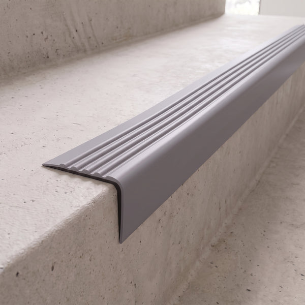 Dark grey 40x40mm flexible PVC angle stair nosing fitted directly to the bare concrete step edge of a daylit stairwell, showing the rounded nose and fluted anti-slip tread