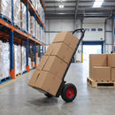 Heavy duty 300kg sack truck loaded with four stacked brown cardboard cartons and tilted back on its pneumatic wheels in a warehouse aisle, with blue and orange pallet racking to the left and an open loading bay door behind