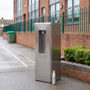 Stainless steel drinking fountain and bottle filling station installed on playground tarmac beside a red brick school wall, green bow-top railings, a shrub planter and a green wheeled bin, with painted court line markings on the ground