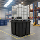 White 1000 litre IBC in a galvanised steel cage standing on a black recycled polyethylene spill pallet inside a warehouse chemical store, with blue steel drums, a yellow hand pallet truck and yellow hazard floor markings around it