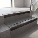 Square-edge aluminium stair nosing with a deep 56mm riser face in Dark Bronze, fitted along two vinyl-covered concrete steps in an office stairwell, with a Mid Grey anti-slip PVC insert running the full width of each step