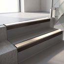 Square-edge aluminium stair nosing with a deep 56mm riser face in Dark Bronze, fitted along two vinyl-covered concrete steps in an office stairwell, with a Sahara anti-slip PVC insert running the full width of each step