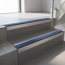 Square-edge aluminium stair nosing with a deep 56mm riser face in Mill Finish, fitted along two vinyl-covered concrete steps in an office stairwell, with a Blue anti-slip PVC insert running the full width of each step