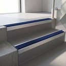 Square-edge aluminium stair nosing with a deep 56mm riser face in Mill Finish, fitted along two vinyl-covered concrete steps in an office stairwell, with a Dark Blue anti-slip PVC insert running the full width of each step