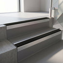 Square-edge aluminium stair nosing with a deep 56mm riser face in Mill Finish, fitted along two vinyl-covered concrete steps in an office stairwell, with a Premium Black anti-slip PVC insert running the full width of each step
