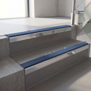 Square-edge aluminium stair nosing with a deep 56mm riser face in Mirroflect Silver, fitted along two vinyl-covered concrete steps in an office stairwell, with a Blue anti-slip PVC insert running the full width of each step