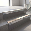 Square-edge aluminium stair nosing with a deep 56mm riser face in Mirroflect Silver, fitted along two vinyl-covered concrete steps in an office stairwell, with a Sahara anti-slip PVC insert running the full width of each step