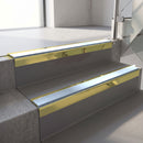 Square-edge aluminium stair nosing with a deep 56mm riser face in Mirrorflect Gold, fitted along two vinyl-covered concrete steps in an office stairwell, with an Aura anti-slip PVC insert running the full width of each step