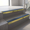 Square-edge aluminium stair nosing with a deep 56mm riser face in Mirrorflect Gold, fitted along two vinyl-covered concrete steps in an office stairwell, with a Blue anti-slip PVC insert running the full width of each step