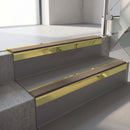 Square-edge aluminium stair nosing with a deep 56mm riser face in Mirrorflect Gold, fitted along two vinyl-covered concrete steps in an office stairwell, with a Brown anti-slip PVC insert running the full width of each step