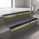 Square-edge aluminium stair nosing with a deep 56mm riser face in Mirrorflect Gold, fitted along two vinyl-covered concrete steps in an office stairwell, with a Dark Blue anti-slip PVC insert running the full width of each step