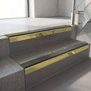 Square-edge aluminium stair nosing with a deep 56mm riser face in Mirrorflect Gold, fitted along two vinyl-covered concrete steps in an office stairwell, with a Dark Grey anti-slip PVC insert running the full width of each step