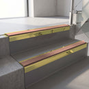Square-edge aluminium stair nosing with a deep 56mm riser face in Mirrorflect Gold, fitted along two vinyl-covered concrete steps in an office stairwell, with a Mid Brown anti-slip PVC insert running the full width of each step