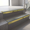 Square-edge aluminium stair nosing with a deep 56mm riser face in Mirrorflect Gold, fitted along two vinyl-covered concrete steps in an office stairwell, with a Mid Grey anti-slip PVC insert running the full width of each step