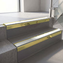 Square-edge aluminium stair nosing with a deep 56mm riser face in Mirrorflect Gold, fitted along two vinyl-covered concrete steps in an office stairwell, with an Oyster anti-slip PVC insert running the full width of each step