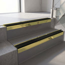 Square-edge aluminium stair nosing with a deep 56mm riser face in Mirrorflect Gold, fitted along two vinyl-covered concrete steps in an office stairwell, with a Premium Black anti-slip PVC insert running the full width of each step
