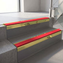 Square-edge aluminium stair nosing with a deep 56mm riser face in Mirrorflect Gold, fitted along two vinyl-covered concrete steps in an office stairwell, with a Red anti-slip PVC insert running the full width of each step