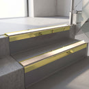 Square-edge aluminium stair nosing with a deep 56mm riser face in Mirrorflect Gold, fitted along two vinyl-covered concrete steps in an office stairwell, with a Sahara anti-slip PVC insert running the full width of each step