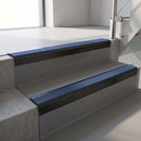Square-edge aluminium stair nosing with a deep 56mm riser face in Satin Black, fitted along two vinyl-covered concrete steps in an office stairwell, with a Blue anti-slip PVC insert running the full width of each step